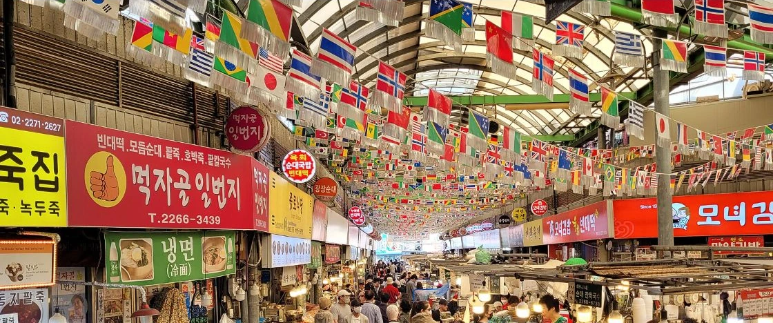 Wide interior view of Gwangjang Market in Seoul with Korean stall signs and a distant public crowd